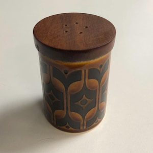 May include: A cylindrical salt shaker with a wooden lid featuring four holes. The ceramic body is brown with a repeating black and brown geometric pattern. The salt shaker has a vintage, retro aesthetic.