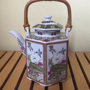 May include: A white ceramic teapot with a hexagonal body and a bamboo handle. The teapot is decorated with floral and bird motifs in shades of pink, green, and blue. The lid is also decorated with floral patterns.