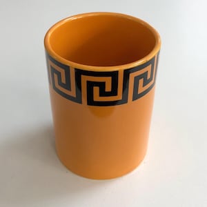 May include: Orange ceramic cylinder with a black Greek key pattern around the top. The cylindrical container is empty and has a smooth, glossy finish. The design is a classic, geometric border.