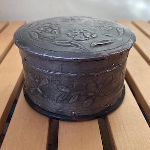 May include: A round, antique metal box with a lid. The lid and sides are decorated with embossed floral and leaf designs. The box is silver-toned with a dark base and sits on a wooden surface.