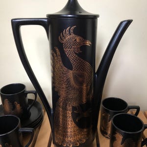 May include: A tall black coffee pot with a lid and a matching set of mugs, all featuring a detailed copper-coloured phoenix illustration. The coffee pot has a long handle and a slender spout, while the mugs are cylindrical with side handles.