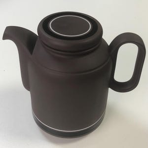 May include: A brown ceramic teapot with a rounded body, a curved spout, and a large handle. The lid has a black and white circular design. A white stripe accents the base. The teapot is designed for brewing tea.