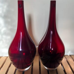 May include: A pair of tall, deep red glass vases with a smooth, teardrop-shaped body and a long, slender neck. Each vase features a clear, thick glass base and a small, circular sticker near the top of the neck, showcasing a translucent finish.