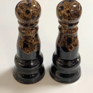 May include: A set of two ceramic salt and pepper shakers. The shakers are black with a brown and black speckled glaze. The shakers are shaped like a tall, slender bottle with a round top.