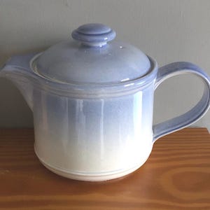 May include: A white and light blue ceramic teapot with a lid and a handle. The teapot has a gradient design with a darker blue at the top and a lighter blue at the bottom.