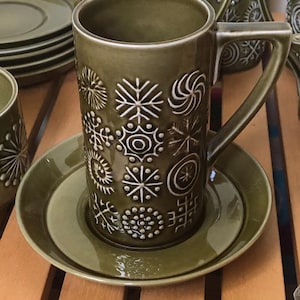 May include: A tall olive green ceramic mug with a matching saucer, featuring a repeating embossed pattern of white geometric star and sunburst motifs. The mug has a straight cylindrical body and a sharp, angular handle, with a glossy finish.
