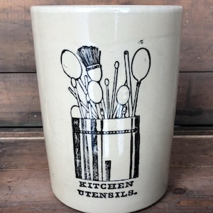 May include: A white ceramic utensil holder with a black and white illustration of kitchen utensils in a container. The text "KITCHEN UTENSILS." is printed on the bottom of the holder.