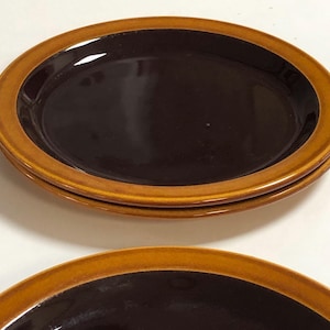 May include: Four oval ceramic plates stacked. Each plate has a dark brown centre and a light brown rim. The plates are stacked, with the top plate at a slight angle, showing the layers.