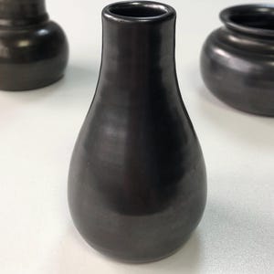 May include: A black ceramic vase with a teardrop shape and a narrow neck. The vase has a smooth, glossy finish and is surrounded by other dark ceramic vessels. The background is a plain white surface.