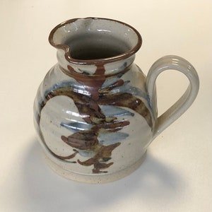 May include: A white ceramic jug with a brown and blue abstract design. The jug has a rounded body and a handle.