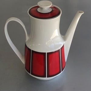 May include: White ceramic coffee pot with a red and black geometric design. The pot has a lid and a spout.