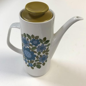 May include: White ceramic coffee pot with a floral design in blue and green. The pot has a curved spout, a handle, and a mustard-coloured lid. A vintage-style coffee server for the kitchen.