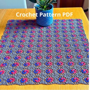 May include: A gray crocheted blanket with a repeating pattern of pink and blue interlocking shapes. The text "Crochet Pattern PDF" is displayed above the blanket. A blue patterned pot with a plant is in the background.