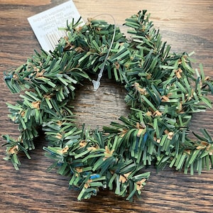 May include: A small, artificial Christmas wreath made of green plastic pine needles with brown accents. A clear plastic tag is attached with the text "MADE IN CHINA" and "For Decorative Use Only".