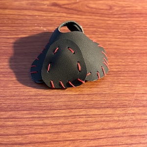 May include: A black leather archery finger tab with red stitching. The tab has a hole for the index finger and is designed to protect the fingers from the string.