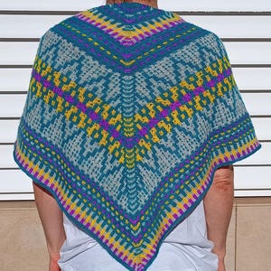 Könnte beinhalten: Ein gestrickter Schal mit geometrischem Muster. Der Schal hat eine türkisfarbene Basis mit gelben, lila und grauen Akzenten. Das Design ist symmetrisch, mit einem zentralen Punkt und strahlenförmigen Mustern. Der Schal wird über einem weißen T-Shirt getragen.