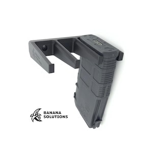 May include: A dark gray magazine holder with the text "BANANA SOLUTIONS" and a banana logo. The holder is designed to hold a magazine and is made of a durable material. The holder is mounted on a white background.