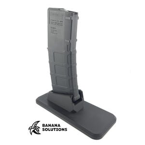 May include: A black magazine holder with a magazine inserted. The holder has a rectangular base and a vertical support for the magazine. The magazine is dark gray and has text printed on it. The logo "BANANA SOLUTIONS" is in the lower left corner.
