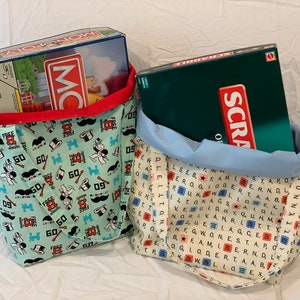 May include: Two fabric tote bags, one with a Monopoly theme and the other with a Scrabble theme. The Monopoly bag is light green with Monopoly characters and the Scrabble bag has a Scrabble tile pattern.