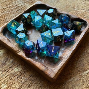 May include: A wooden tray filled with blue and green iridescent dice with gold numbers. The dice are in a variety of shapes and sizes.