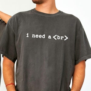 May include: Two people wearing gray t-shirts with the text "I need a <br>" printed on the front. The shirts are a relaxed fit and have a vintage look.