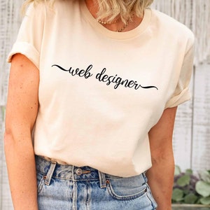 May include: A cream-colored t-shirt with the text "web designer" in a black script. The shirt is paired with light blue jeans. The word "Natural" is in a light blue circle in the upper left corner.