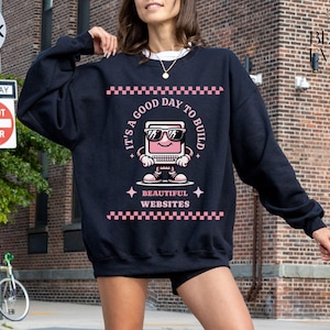 Op de afbeelding: Zwart sweatshirt met een roze en witte afbeelding van een computer met de tekst "Het is een goede dag om mooie websites te bouwen".