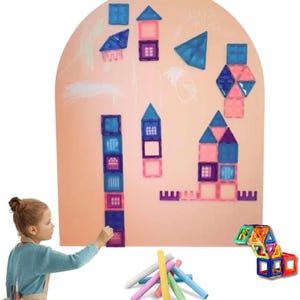 Op de afbeelding: Een kind gebruikt kleurrijke krijtjes om te tekenen op een groot, gebogen magnetisch whiteboard. Het bord is bedekt met kleurrijke magnetische tegels in de vorm van huizen, sterren en geometrische vormen. Een set kleurrijke krijtjes ligt op de voorgrond.