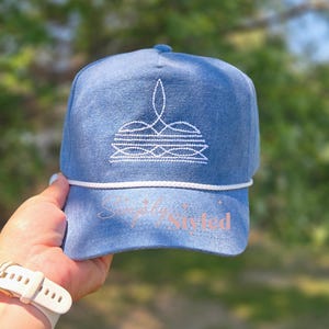May include: A blue denim baseball cap featuring a white embroidered crown-like design on the front panel. A white braided cord runs across the base of the crown. The text Simply Styled is printed in a light pink color on the curved brim of the cap.