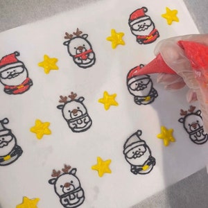 May include: A white surface with a pattern of outlined Christmas characters and stars. The characters include Father Christmas and reindeer. The stars are yellow. A red piping bag is being used to fill in the characters with red icing.
