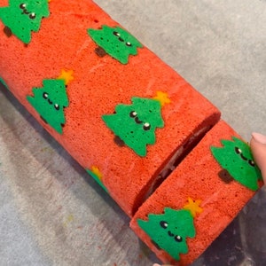 以下が含まれることがあります： 白の詰め物が入った赤と緑のクリスマスのロールケーキ。ロールケーキは、黄色の星と黒い目で飾られた緑のクリスマスツリーで飾られています。ロールケーキはナイフで切られています。
