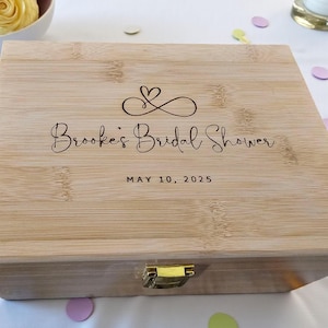 May include: A light-colored wooden box with a hinged lid and a gold-colored clasp. The box is engraved with the words "Brooke's Bridal Shower" and the date "MAY 10, 2025", along with a heart and infinity symbol design.