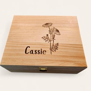 May include: A light-colored wooden box with a floral design and the name "Cassie" engraved on the lid. The box has a rectangular shape and a gold-colored clasp.