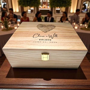 May include: A light-colored wooden box with a gold clasp. The lid features a heart made of fingerprints, the names "Clara + Will", the word "GOLDEN", and the date "JUNE 11, 2025". The box is on a dark brown table.