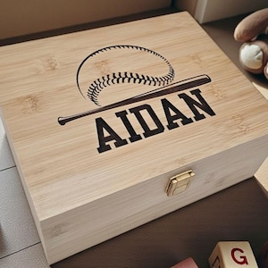 May include: A rectangular light-colored wooden box with a hinged lid and a gold-tone metal latch. The lid features a laser-engraved graphic of a baseball and a baseball bat positioned above the name AIDAN in bold, dark, capital letters.