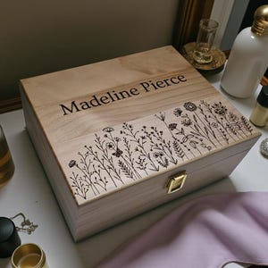 May include: A rectangular light-colored wooden box with a hinged lid featuring a gold-tone metal latch. The lid is engraved with the name Madeline Pierce and a detailed black line art illustration of various wildflowers and stems.