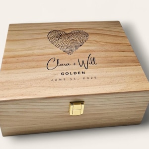 May include: A wooden box with a gold clasp. The lid features a heart-shaped fingerprint design and the names "Clara + Will" with the word "GOLDEN" and the date "JUNE 11, 2025" engraved on it.