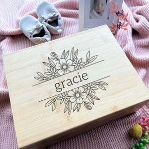 May include: A rectangular light-colored bamboo wooden box featuring a laser-engraved design of flowers and leaves surrounding the name gracie in lowercase letters. The box has a smooth finish and a hinged lid, showcasing the natural wood grain.