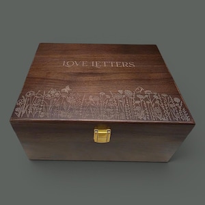May include: A wooden box with the words "LOVE LETTERS" engraved on the lid. The box features a floral design along the edge and a gold-colored clasp. The box is a dark brown color.