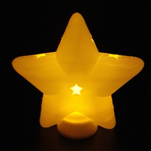 May include: A glowing, star-shaped night light with a small star cutout in the center. The light emits a warm yellow glow against a black background. The star is approximately 5 inches wide.