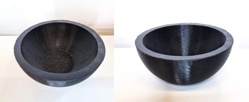 May include: A dark gray, semi-spherical bowl with a textured interior and a smooth, rounded rim. The bowl is made of a solid material and sits on a white surface. The design is simple and modern, suitable for various uses.