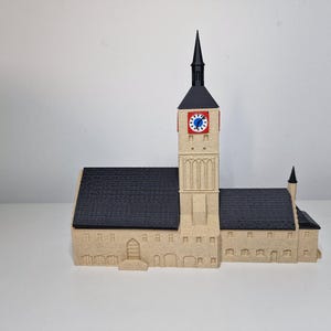 May include: A beige model of a church with a tall clock tower and a black steeple. The clock face is red with blue and white details. The roof is black, and the building has multiple windows.