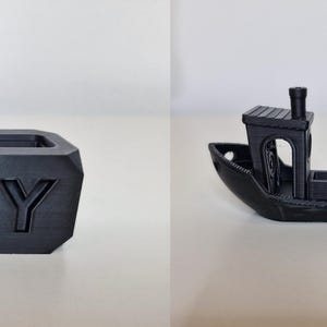 May include: Two black 3D-printed objects: a small boat and a cube. The boat has a cabin and smokestack. The cube has the letter "Y" on one side. Both objects are on a white surface.