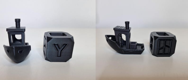 May include: Two black 3D-printed objects: a small boat and a cube. The boat has a cabin and smokestack. The cube has the letter "Y" on one side. Both objects are on a white surface.