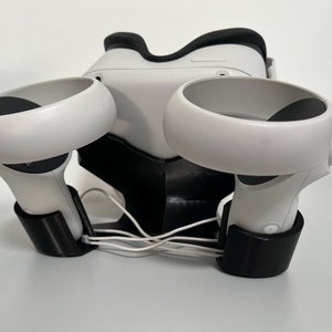 May include: A white VR headset and controllers are displayed with a black stand. The controllers are positioned on either side of the headset, with white cables neatly arranged. The stand is black and holds the VR equipment.