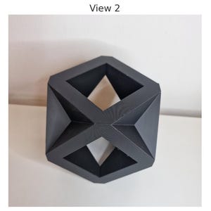 May include: A dark gray geometric sculpture with a multifaceted design. The sculpture has open spaces and angular shapes, creating a modern and abstract aesthetic. The object is displayed on a white surface.