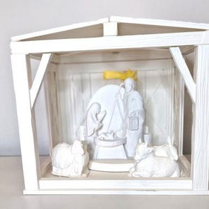 May include: A white wooden nativity scene with a pitched roof and a sliding door. Inside, white figurines depict the holy family and animals. The scene is set inside a small, open-fronted structure.