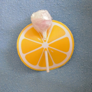 Op de afbeelding: Een gele citroenschijf-lolly met een roze en witte lolly erop.