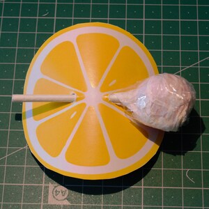 Op de afbeelding: Een gele papieren citroenschijf met een witte stok en een roze suikerspinlolly die in het midden is bevestigd.