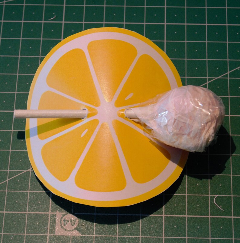 Op de afbeelding: Een gele papieren citroenschijf met een witte stok en een roze suikerspinlolly die in het midden is bevestigd.